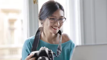 Cheerful woman with camera using laptop. Andrea Piacquadio via Pexels